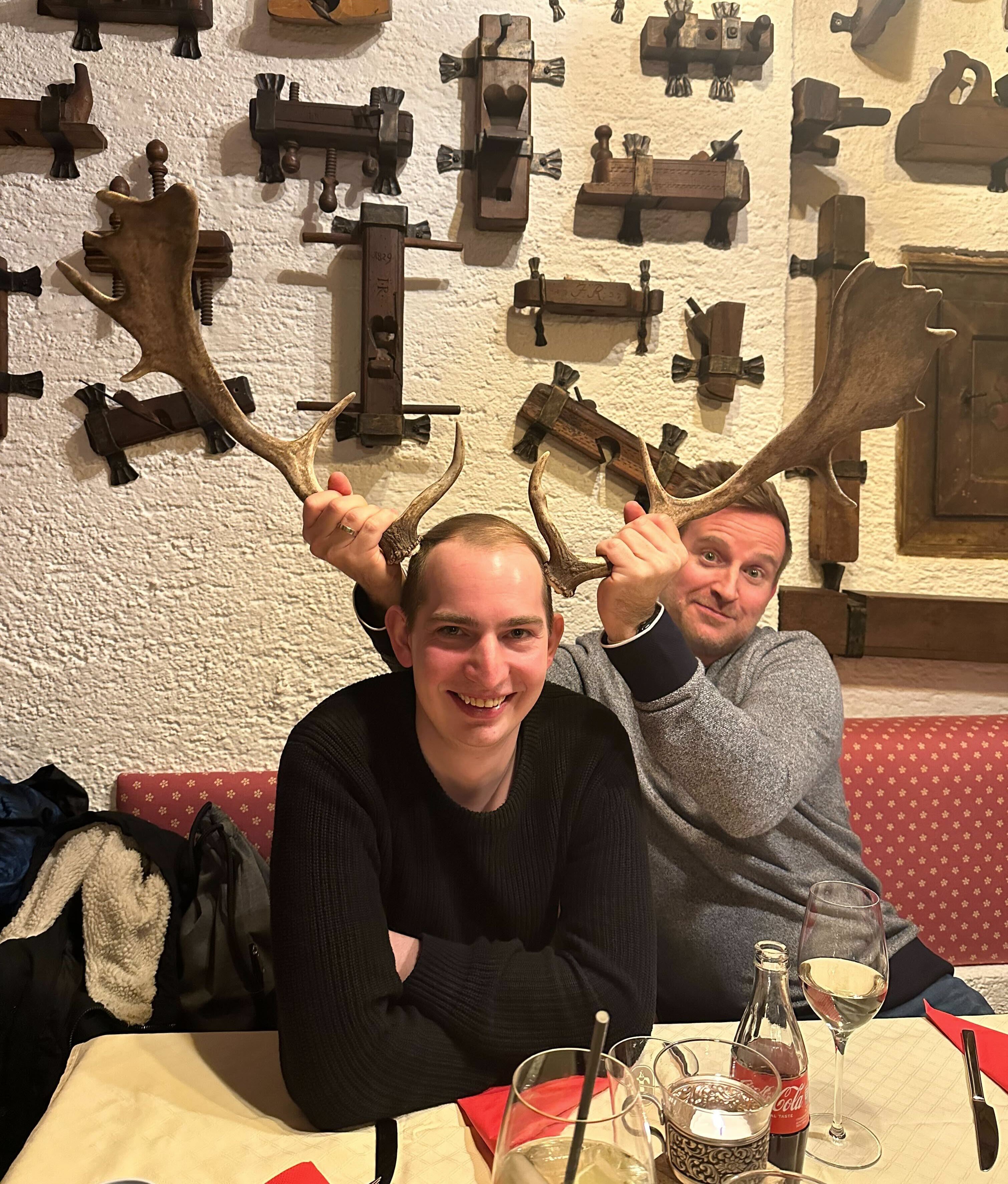 Zwei Männer scherzen im Wirtshaus: Einer hält Hirschgeweihe über den Kopf des anderen. Stube mit alten Holzwerkzeugen an der Wand, Getränke am Tisch.